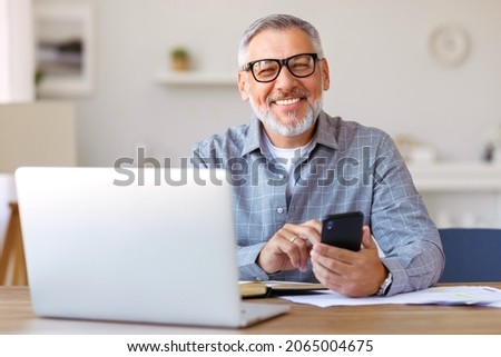 Handsome smiling senior man wearing glasses using mobile phone while sitting at his cozy workplace with laptop at home, retired male chatting with friends in social media, typing on smartphone Royalty-Free Stock Photo #2065004675