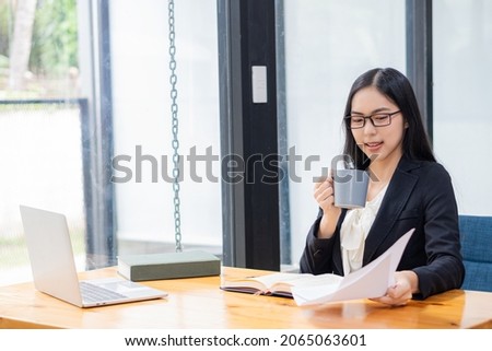Serious woman sitting at a desk and working on laptop from home. Smiling Asian woman working, reading papers, report on successful female freelancer project. use a computer	