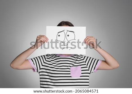Woman hiding behind sheet of paper with sad face on grey background