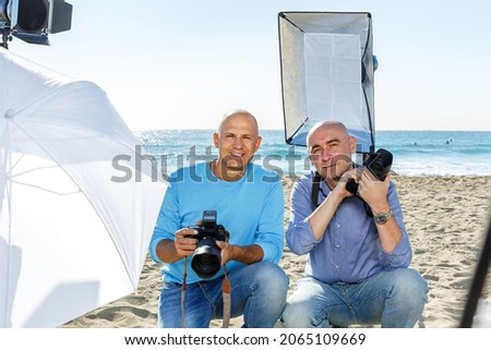 Portrait of two smiling photographers with cameras among professional photo equipment on seacoast
