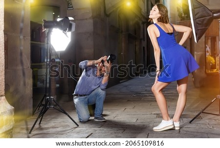 Photographer using professional camera and light equipment for taking pictures of adult woman in blue dress on town street