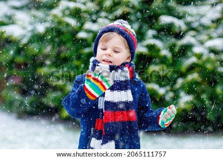Cute little funny child in colorful winter fashion clothes having fun and playing with snow, outdoors during snowfall. Active outdoors leisure with children. Kid boy and toddler catching snowflakes.