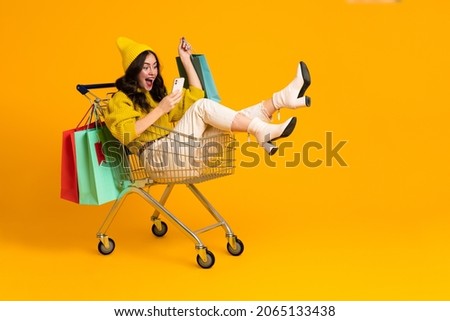 White excited woman laughing and using cellphone in shopping cart isolated over yellow background Royalty-Free Stock Photo #2065133438