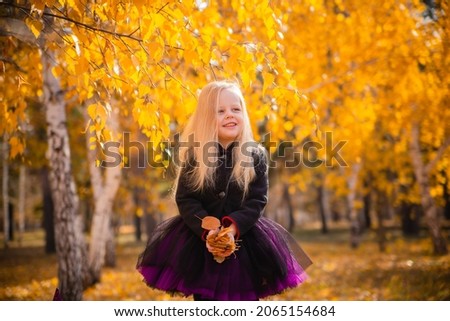 Little cute blonde girl dressed as a witch throws leaves in the park in autumn and smiles. Decorations for All Saints Day. Halloween costume. Child rejoice