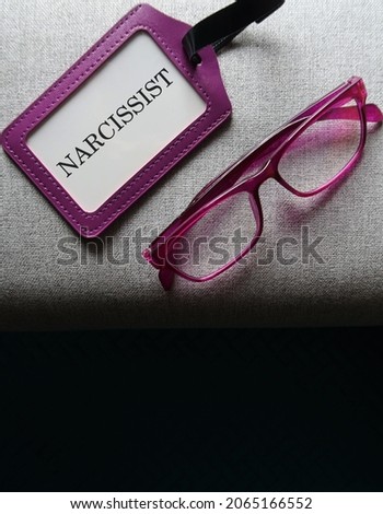 Purple reading glasses and ID card on black copy space and text NARCISSIST, means self-centered arrogant thinking and behavior, people who lack of empathy for others, excessive need of admiration  Royalty-Free Stock Photo #2065166552