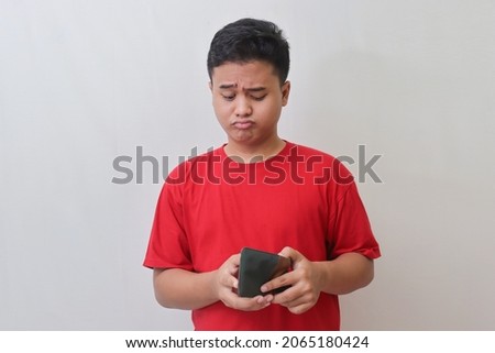 Portrait of sad Asian man in red t-shirt looking at his empty wallet. Isolated image on gray background Royalty-Free Stock Photo #2065180424