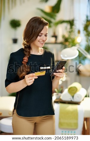 smiling 40 years old woman employee with smartphone and credit card buying online in modern beauty salon.