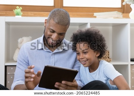 Caring young african american father using touchpad gadget with small adorable kid son, enjoying playing games online, watching funny cartoons or shopping in internet store, tech addiction concept.