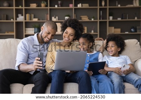 Happy young african american couple parents and cute little children using different gadgets, spending time online, playing games, entertaining sitting together on sofa in living room, tech addiction.