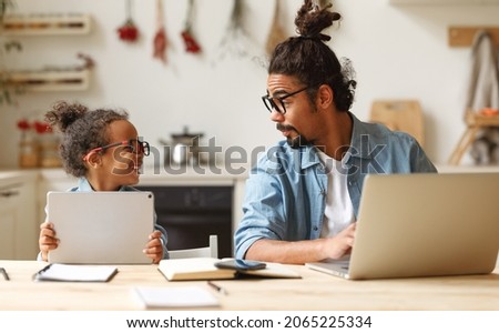 Young african american man father working remotely on laptop computer while his happy little son using digital tablet, sitting together and wooden table at home, dad trying to work online with kids