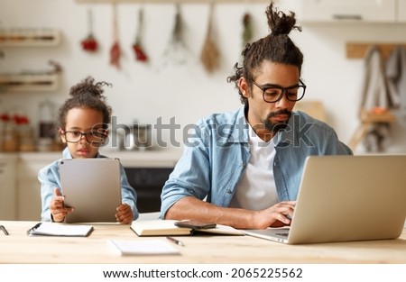 African american father and son using gadgets laptop and tablet while sitting at desk at home, dad working on computer while his kid little boy playing games or browsing internet on digital device