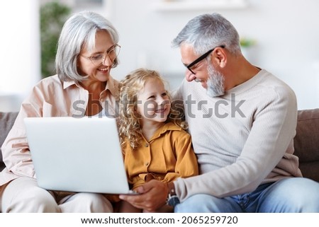 Smiling senior grandparents and cute little girl granddaughter looking at laptop computer screen while sitting on sofa at home, happy child watching cartoons with grandpa and grandma on weekend
