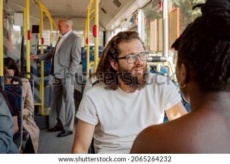 Young man with thick beard long hair wearing glasses sits on public transport bus in front of dark-skinned colleague with dreadlocks tied up in bun friends returning from work college talking laughing Royalty-Free Stock Photo #2065264232