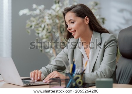 Focused on work attractive pretty brunette girl in classic makeup dressed in shirt and jacket, sits at a desk in the company in front of laptop, the secretary reads email, writes back messages smiles