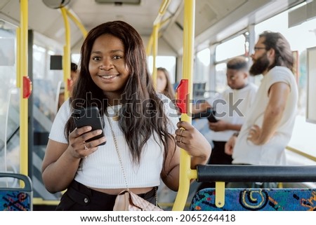A dark-skinned woman rides in a public transport bus holding on to a barrier, a girl buys an electronic ticket on her phone through an app, other passengers in the vehicle in the background