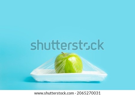 Fruits in plastic packaging from the supermarket are minimal. Apple in cellophane and non-degradable plastic on a blue background. Biodegradable product packaging, environmental protection, nature Royalty-Free Stock Photo #2065270031