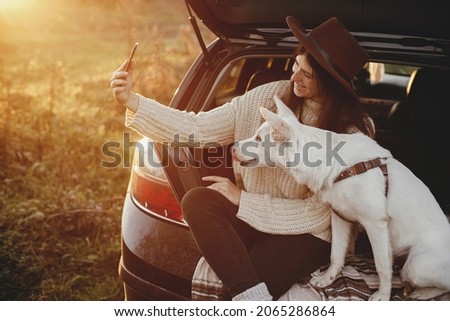 Stylish hipster woman taking selfie photo with cute dog in car trunk in warm sunset light. Autumn road trip with pet. Young female in hat and sweater using phone and travelling with sweet white dog
