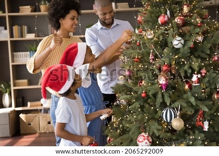 Happy young african american parents decorating fir Christmas tree with adorable little kids, putting lights toys on fluffy branches, enjoying preparing for New Year winter holidays celebration.