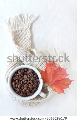 a cup with roasted coffee beans in a knitted light scarf decorated with a maple leaf top view. autumn warming aromas