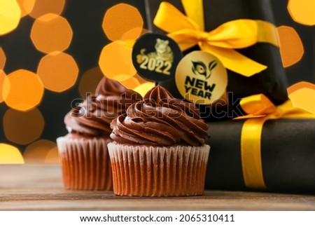 Tasty cupcakes for New Year 2022 celebration with gifts on table against blurred lights
