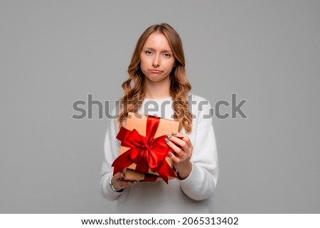 Young woman in white sweater holding gift box looking at camera with displeased upset expression, standing over gray background