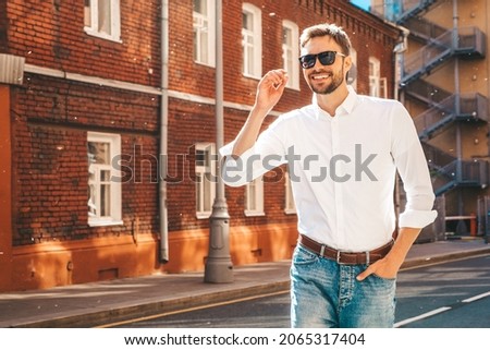 Portrait of handsome confident stylish hipster lambersexual model.Modern man dressed in white shirt. Fashion male posing on the street background in sunglasses. Outdoors at sunset 