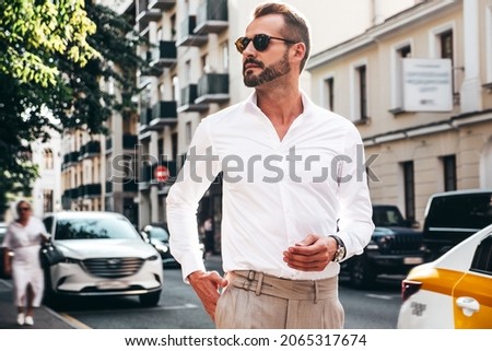 Portrait of handsome confident stylish hipster lambersexual model.Sexy modern man dressed in white shirt and trousers. Fashion male posing on street background in Europe city at sunset. In sunglasses Royalty-Free Stock Photo #2065317674