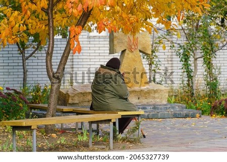 An old woman prays on a bench under an autumn tree near a stone cross on the territory of a Christian church 