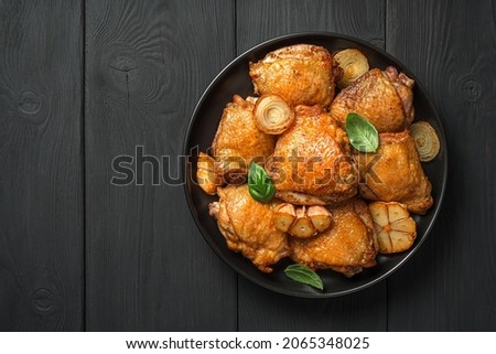 Fried chicken thighs with garlic, onion and basil in a black plate on a black background. Top view, copy space.