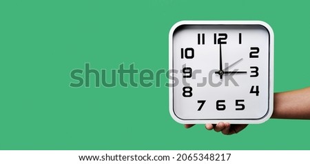 a young man holding a clock being set backward or forward, at the end or beginning of the summer time, on a green background, in a panoramic format to use as web banner or header Royalty-Free Stock Photo #2065348217