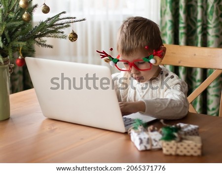  Little boy in a white sweater and funny glasses sits at a laptop and writes a letter to Santa Claus.