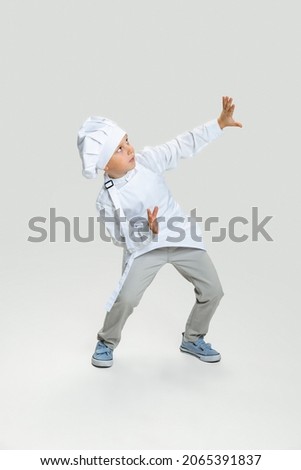 Looks scared. Cute little boy in white cook uniform and huge chef's hat isolated on white studio background. Concept of childhood, studying, jobs, games, emotions. Copy space for ad, text