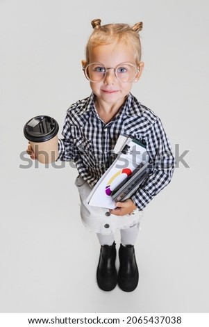 Little worker. Cute playful girl, child with coffee cup and notes isolated on gray background. Model wearing glasses and checkered shirt. Concept of childhood, education, motherhood. Copy space for ad