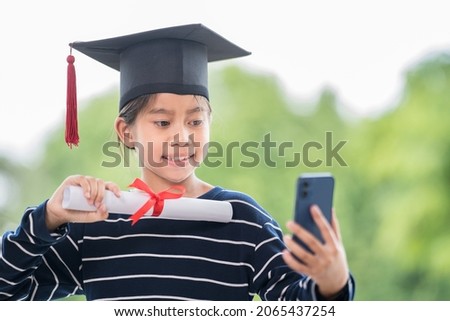 Happy Asian female children graduate with graduation cap and rolled graduation certificate holding smartphone to call and chat with friends and family. Education Concept Stock Photo