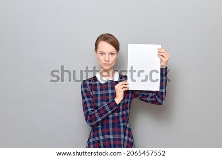 Studio portrait of serious focused young blond woman wearing checkered dress, holding white blank paper sheet with place for your text and design in her hands, standing alone over gray background