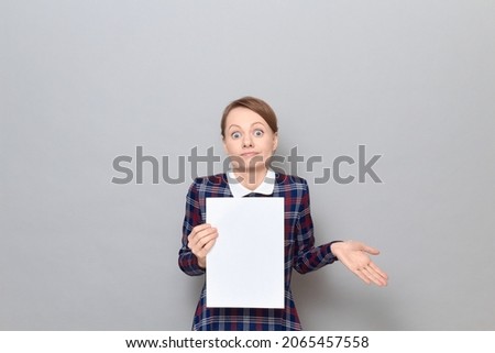 Studio portrait of funny confused young blond woman holding white blank paper sheet with place for your text, shrugging her shoulders, looking bewildered and silly, standing alone over gray background