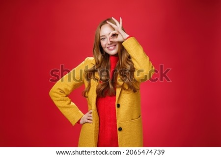 Bright and sunny girl feeling happy even on rainy days standing in yellow stylish coat over red background showing okay gesture over eye, smiling broadly at camera, holding hand on waist