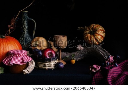 A wooden wine glass surrounded by a large tree branch, pumpkins, a jar of jam, a jug of wine, apples, plums and branches of wild grapes. Low key. Still-life. Horizontal picture.