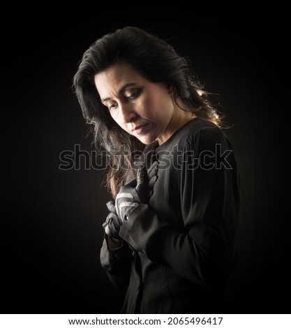 Brunette woman in black standing in front of a black backdrop, clasping her hands to her chest and looking down with a serious, sad expression on her face