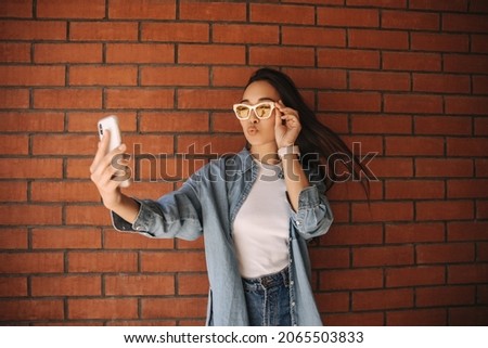 Fashionable young asian teenager makes selfie sending kiss to camera against background of wall. Her dark long hair flutters in wind, holding glasses on face with her hand.