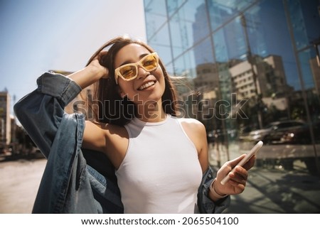 Close-up of fashionably dressed asian tourist in stylish glasses, eyes closed, walking relaxed in center of capital. Brunette in denim jacket with phone in hand. Lifestyle, female beauty concept Royalty-Free Stock Photo #2065504100