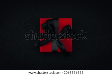 One red small gift box with a black ribbon tied in a bow lies in the middle on a black background, close-up top view. Christmas and black friday concept.