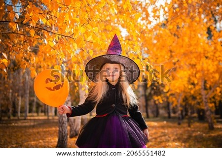 Little cute blonde girl dressed as a witch holding an orange balloon in the park in autumn. Decorations for All Saints Day. Halloween costume. Child in a sorceress hat