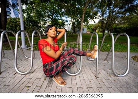 Photo of a beautiful female model posing outdoors in a park scene. Woman is posing by a metal bike rack in the park. 
