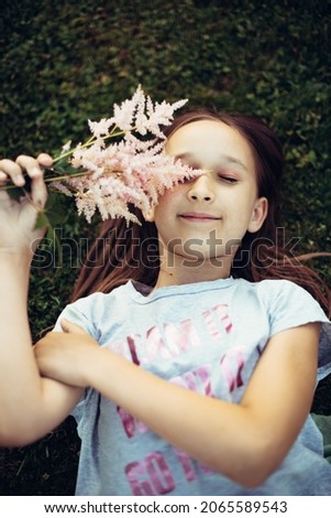 Photo from above of a little girl with long dark hair posing with flowers in her hands lying on the grass. Portrait of a child with flowers outdoors. Selective focus, defocus, toning.