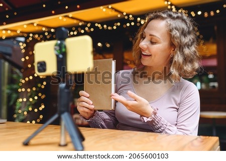 Young blogger fun woman 20s wear casual clothes sit alone at table in cafe shop take picture on mobile cell phone do selfie shot photo with book point finger on textbook relaxing in restaurant indoors