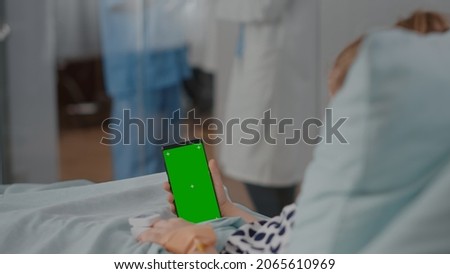 Hospitalized little child resting in bed looking at mock up green screen chroma key smartphone with isolated display. Sick kid suffering medical surgery during recovery consultation in hospital ward