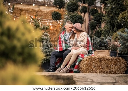 A beautiful young couple in white clothes are sitting near a Christmas tree in the garden. Happy man and woman, romance, Christmas celebration, fun, love.