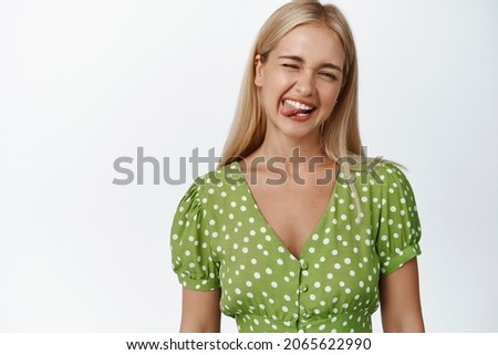 Funny blond woman shows tongue and smiles, winks at camera happy, stands against white background in green dress