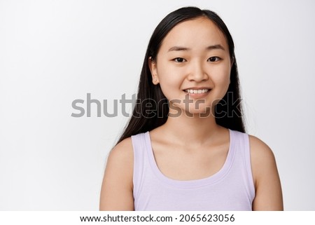 Close up portrait of attractive aisan woman with white smile, looking happy, standing in tank top over white background. Skin care concept
